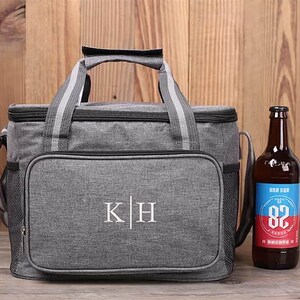 Op de afbeelding: Een grijze ge&iuml;soleerde koeltas met een zwarte rits en een zwarte handgreep. De tas heeft een wit geborduurd monogram "K|H" aan de voorkant. Naast de tas staat een bruine glazen fles bier.