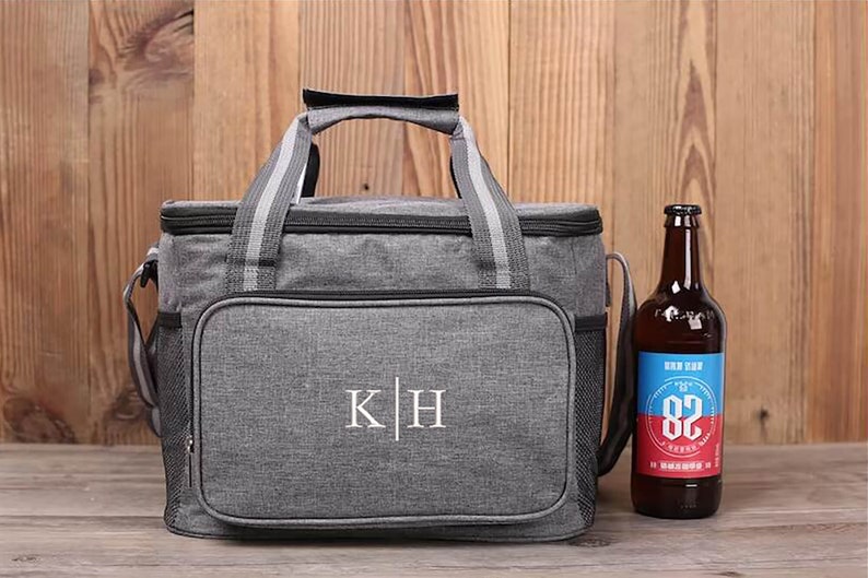 Op de afbeelding: Een grijze ge&iuml;soleerde koeltas met een zwarte rits en een zwarte handgreep. De tas heeft een wit geborduurd monogram "K|H" aan de voorkant. Naast de tas staat een bruine glazen fles bier.