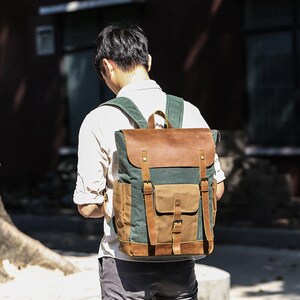 May include: A green and brown canvas backpack with leather accents and buckles. The backpack has a large main compartment and a smaller front pocket.