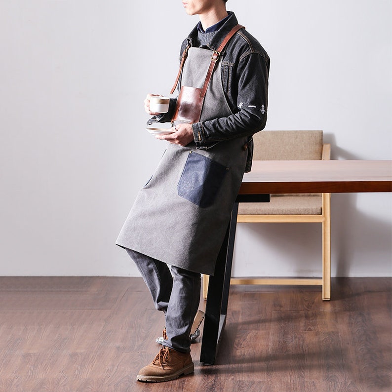 May include: Gray apron with brown leather straps and a brown leather chest pocket. The apron has a large blue pocket on the lower right side. The person is wearing a denim jacket and holding a cup and saucer.