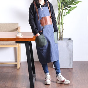 May include: A blue apron with brown leather accents and straps. The apron has two large pockets, one green and one blue, and a brown leather chest pocket. The apron is worn over a black hoodie and blue jeans.