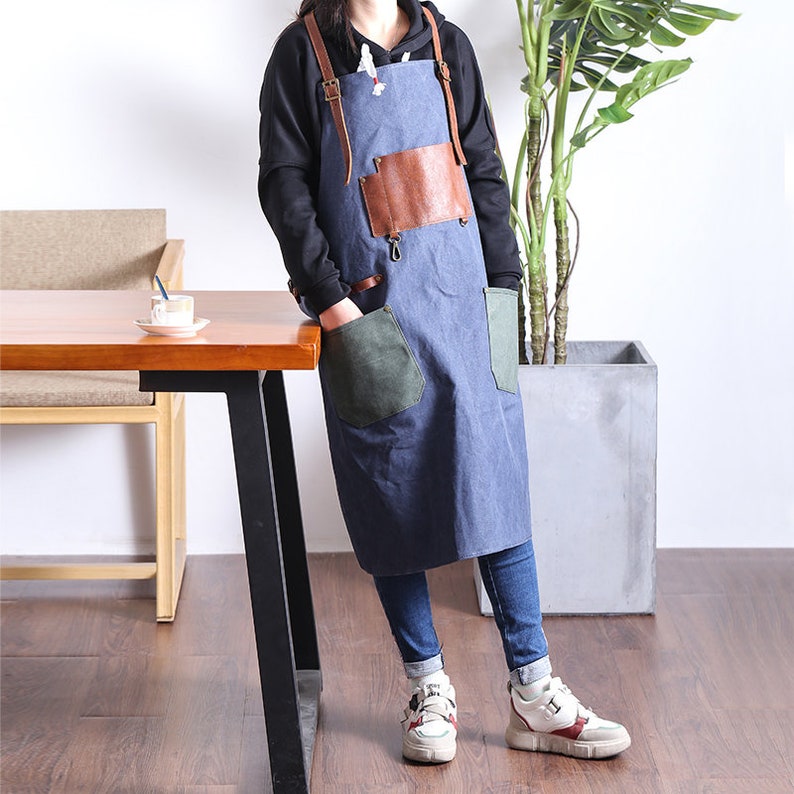 May include: A blue apron with brown leather accents and straps. The apron has two large pockets, one green and one blue, and a brown leather chest pocket. The apron is worn over a black hoodie and blue jeans.