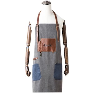 May include: A gray apron with brown leather accents and straps. The apron has two blue pockets and a brown leather patch with the name "Jack" printed on it. The apron is displayed on a mannequin.