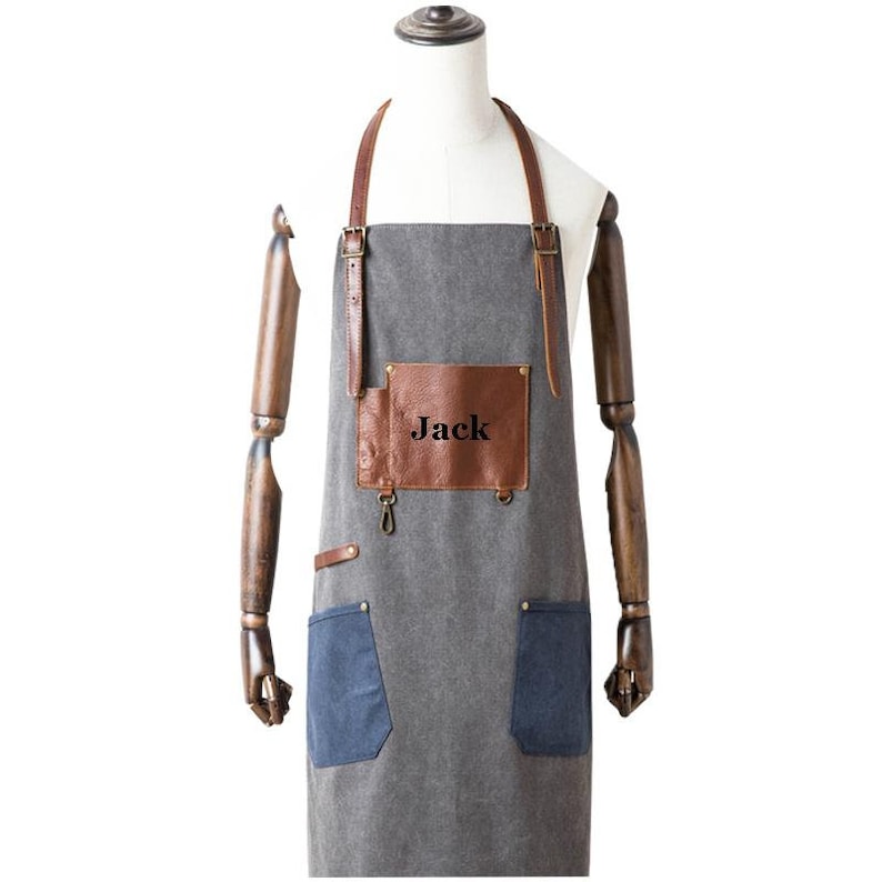 May include: A gray apron with brown leather accents and straps. The apron has two blue pockets and a brown leather patch with the name "Jack" printed on it. The apron is displayed on a mannequin.