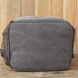 Puede incluir: Una bolsa de almuerzo de lona gris con un bolsillo lateral de malla y un asa de cuero marr&oacute;n.