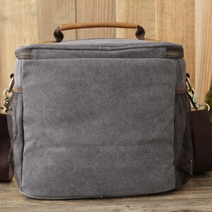 Puede incluir: Una bolsa de refrigeraci&oacute;n de lona gris con un asa y una correa de hombro de cuero marr&oacute;n. La bolsa tiene un bolsillo lateral de malla y un cierre de cremallera. La bolsa est&aacute; sobre una superficie de madera junto a una botella de cerveza Heineken.