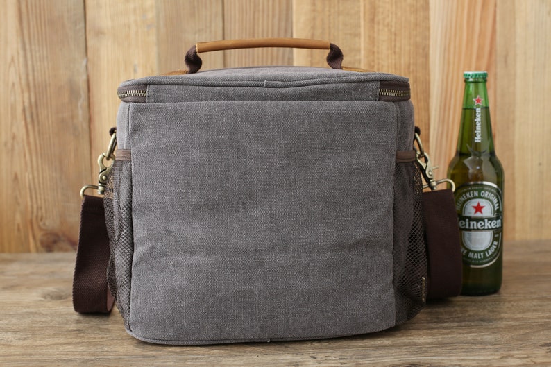 Puede incluir: Una bolsa de refrigeraci&oacute;n de lona gris con un asa y una correa de hombro de cuero marr&oacute;n. La bolsa tiene un bolsillo lateral de malla y un cierre de cremallera. La bolsa est&aacute; sobre una superficie de madera junto a una botella de cerveza Heineken.