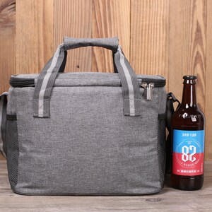 Op de afbeelding: Een grijze ge&iuml;soleerde lunchtas met een ritssluiting en een zwarte schouderband. De tas heeft een zijvak van gaas en staat op een houten oppervlak naast een bruine glazen fles met een blauw en rood etiket waarop "Hop Shop 82 Brewery" staat.