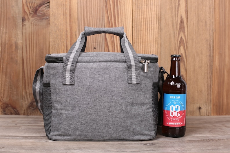 Op de afbeelding: Een grijze ge&iuml;soleerde lunchtas met een ritssluiting en een zwarte schouderband. De tas heeft een zijvak van gaas en staat op een houten oppervlak naast een bruine glazen fles met een blauw en rood etiket waarop "Hop Shop 82 Brewery" staat.