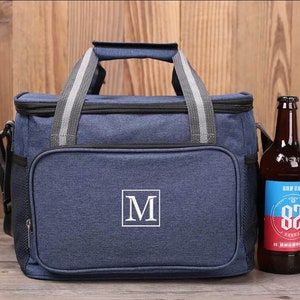 Op de afbeelding: Een blauwe ge&iuml;soleerde koeltas met een ritsvak en een wit monogram "M" aan de voorkant. De tas heeft een bovenste handvat en een schouderband. Naast de tas staat een bruine glazen fles met een etiket.
