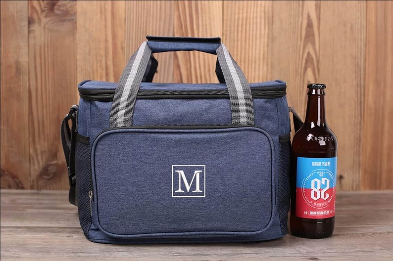 Op de afbeelding: Een blauwe ge&iuml;soleerde koeltas met een ritsvak en een wit monogram "M" aan de voorkant. De tas heeft een bovenste handvat en een schouderband. Naast de tas staat een bruine glazen fles met een etiket.