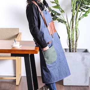 May include: Blue denim apron with brown leather accents, including straps and a chest patch. Features a large green pocket and metal hardware. The apron is designed for kitchen or workshop use.