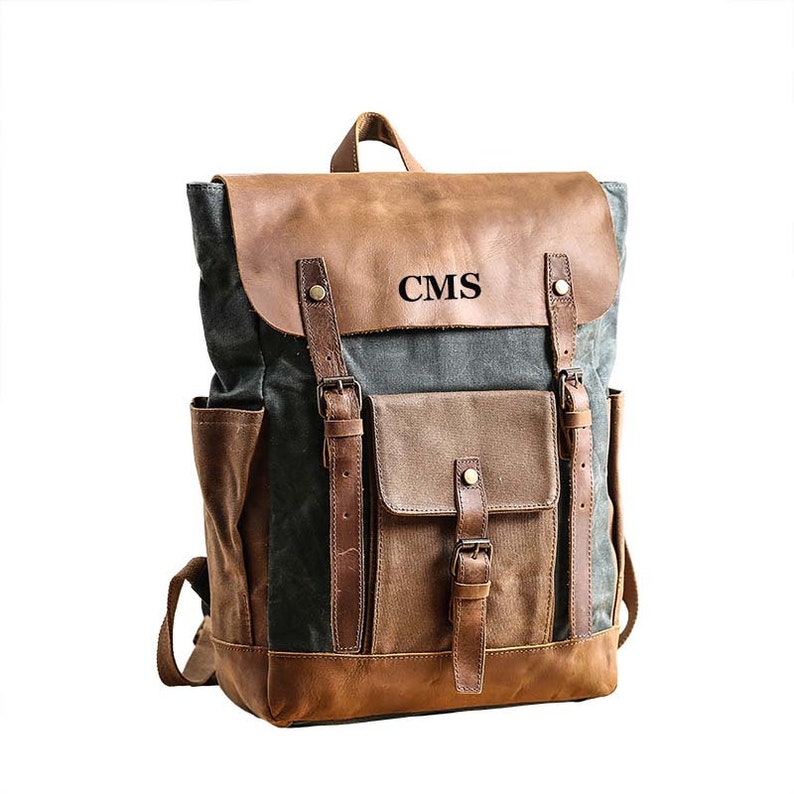 May include: A brown leather and canvas backpack with a large front pocket and adjustable straps. The backpack has the initials "CMS" embroidered on the front.