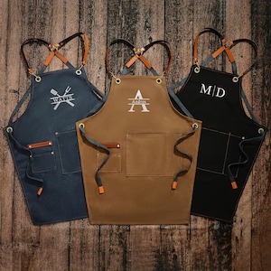 Puede incluir: Tres delantales de lona personalizados con correas ajustables. Los delantales son de color azul, marrón y negro. El delantal azul tiene el texto "WATTS" y un diseño de tenedor y cuchara cruzados. El delantal marrón tiene el texto "Andres" y una letra "A". El delantal negro tiene el texto "M/D".