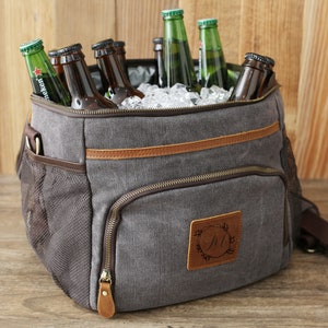 Puede incluir: Una bolsa de enfriador de lona gris con detalles de cuero marr&oacute;n y cierre de cremallera. La bolsa tiene un bolsillo de malla en el lateral y un parche de cuero personalizado con la inicial "M" en un dise&ntilde;o de corona. El enfriador est&aacute; lleno de hielo y botellas de cerveza.