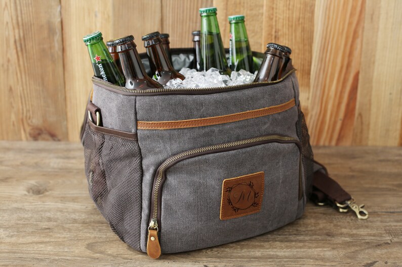 Puede incluir: Una bolsa de enfriador de lona gris con detalles de cuero marr&oacute;n y cierre de cremallera. La bolsa tiene un bolsillo de malla en el lateral y un parche de cuero personalizado con la inicial "M" en un dise&ntilde;o de corona. El enfriador est&aacute; lleno de hielo y botellas de cerveza.