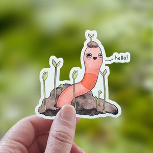 May include: A pink worm with a brown hat and a smiling face is peeking out of the ground.  The worm is surrounded by green shoots and the text "hello!" is written above the worm.