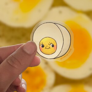 Könnte beinhalten: Ein transparenter Aufkleber mit einer Cartoon-Illustration eines lächelnden, gelben Eies mit einer weißen Schale. Der Aufkleber trägt den Text "pandaspinkle" in der rechten unteren Ecke.