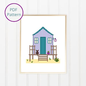 May include: A cross-stitch pattern of a beach house with a striped purple and blue exterior, a brown cat sitting on the porch, and a small potted plant. The house has a teal door and a brown wooden porch with steps.