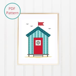 Op de afbeelding: Een ingelijst kruissteekpatroon van een strandhuisje. Het huisje is lichtblauw met witte verticale strepen en een rode deur. Een rood-witte stippen vlag wappert vanaf het dak. De woorden "PDF Pattern" staan in een cirkel linksboven.