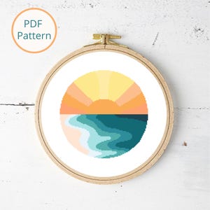 May include: A cross-stitch pattern in a wooden embroidery hoop. The design features a stylized sunset over the ocean, with yellow, orange, and blue hues. The text "PDF Pattern" is in a circle at the top left.