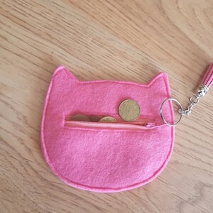 May include: Pink felt cat-shaped coin purse with a zipper closure and a silver key ring. The purse is filled with coins.