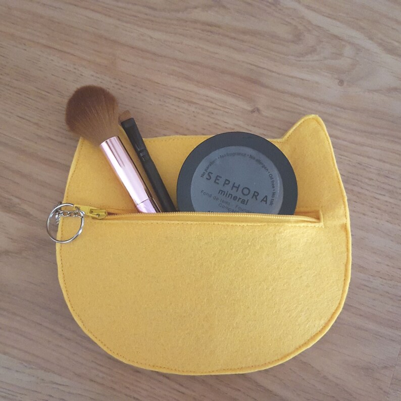 May include: Yellow felt cat-shaped makeup bag with a zipper closure. The bag is open and contains a black makeup compact with the text "SEPHORA mineral" and two makeup brushes.
