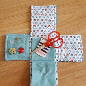 Op de afbeelding: Een blauwe en witte stoffen naaiset met een rode en witte gepatroneerde klep. De set bevat een rood schaartje, naalden, draad en knopen.