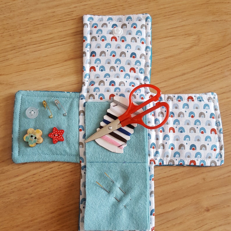 Op de afbeelding: Een blauwe en witte stoffen naaiset met een rode en witte gepatroneerde klep. De set bevat een rood schaartje, naalden, draad en knopen.