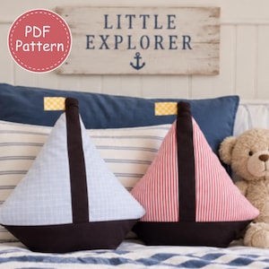 May include: Two triangular sailboat-shaped pillows featuring a dark brown base and mast. One pillow has a light blue grid pattern, and the other has a red and white vertical stripe pattern. Each features a small yellow and white checkered flag at the top.