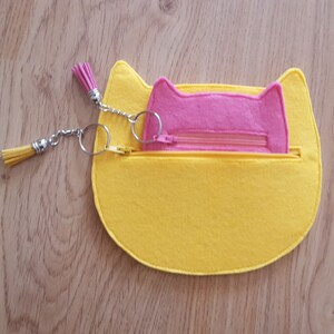 May include: A yellow felt cat-shaped pouch with a pink felt pocket inside. The pouch has a zipper closure and a silver chain with two tassels.