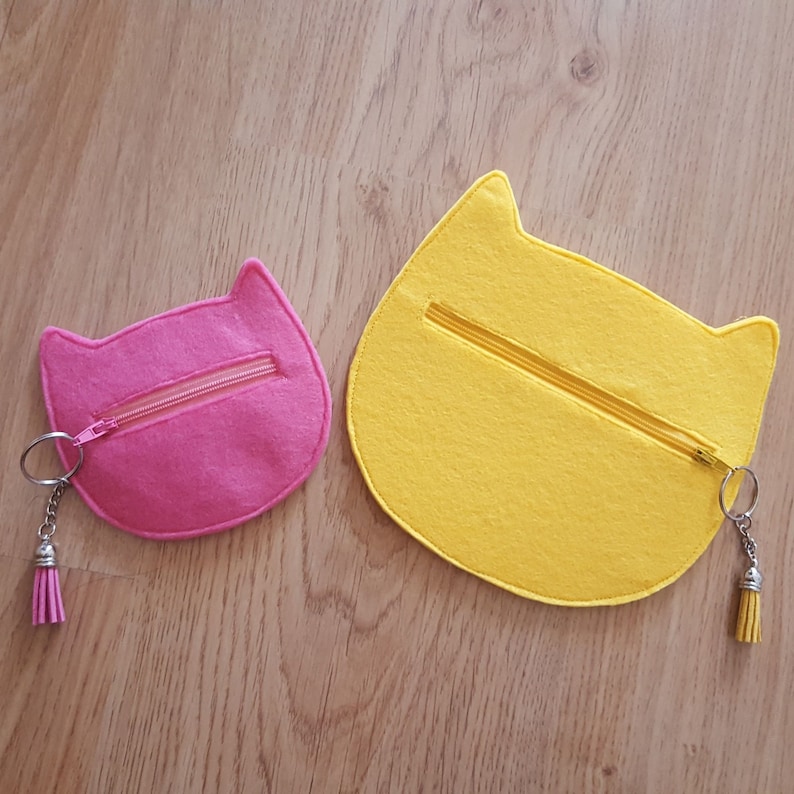 May include: Two felt cat-shaped coin purses with zippers. The smaller purse is pink and the larger purse is yellow.