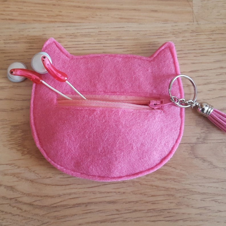 May include: A pink felt cat-shaped pouch with a zipper closure and a silver key chain with a pink tassel. The pouch has two white and red earbuds inside.