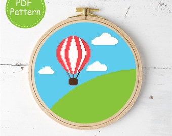 May include: A circular wooden embroidery hoop containing a cross-stitch pattern of a red and white striped hot air balloon floating in a light blue sky with white clouds above a green grassy hill. A green circular badge reads PDF Pattern.
