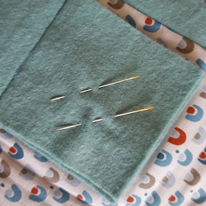 Op de afbeelding: Een close-up van een teal viltstof met vier naalden erin gestoken. De naalden zijn zilver en goud van kleur.