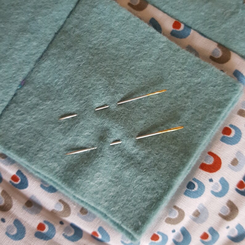Op de afbeelding: Een close-up van een teal viltstof met vier naalden erin gestoken. De naalden zijn zilver en goud van kleur.