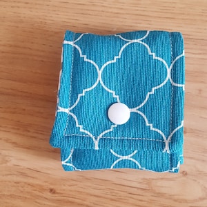 Op de afbeelding: Een klein turquoise blauw stoffen tasje met een wit geometrisch patroon en een witte drukknoop sluiting.