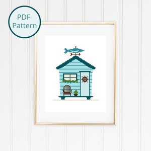 May include: A cross stitch pattern of a blue beach house with a fish weathervane on top. The house has a door with a nautical wheel and a window. There is a chair and a cactus in front of the house.