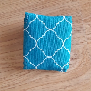 Op de afbeelding: Een kleine, turquoise blauwe stoffen tas met een wit geometrisch patroon. De tas is gemaakt van een zachte, gestructureerde stof en heeft een eenvoudige, rechthoekige vorm.