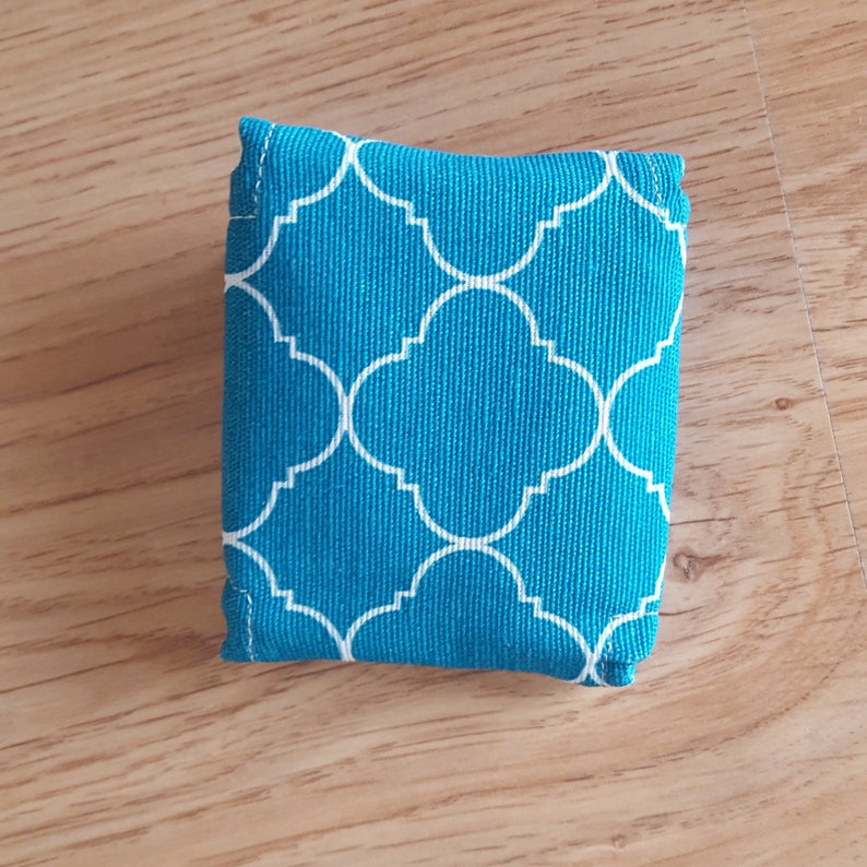 Op de afbeelding: Een kleine, turquoise blauwe stoffen tas met een wit geometrisch patroon. De tas is gemaakt van een zachte, gestructureerde stof en heeft een eenvoudige, rechthoekige vorm.