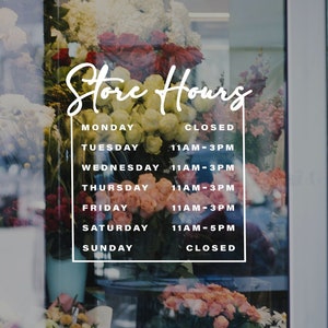 May include: A white sign with black text on a glass door with flowers behind it. The sign says "Store Hours" and lists the hours of operation for each day of the week.  Monday and Sunday are closed.  Tuesday through Friday are open from 11 AM to 3 PM.  Saturday is open from 11 AM to 5 PM.