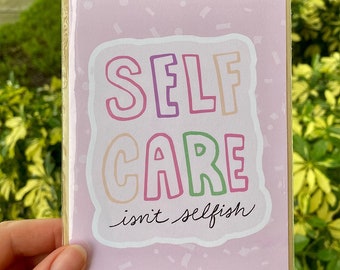 Könnte beinhalten: Eine Grußkarte mit den Worten "SELF CARE isn't selfish" in bunten Buchstaben. Die Karte hat einen rosa Hintergrund mit Konfetti und befindet sich in einer transparenten Plastikhülle.