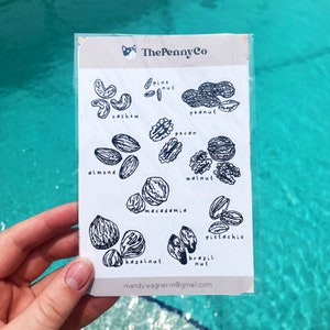 May include: A clear sticker sheet featuring black and white illustrations of various nuts, including cashews, almonds, and walnuts. The text "The Penny Co" is at the top, and the email address "mandy.wagner.m@gmail.com" is at the bottom.