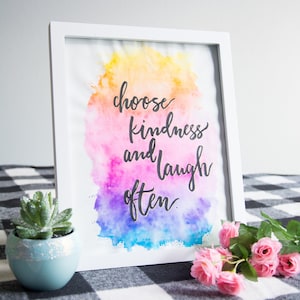 May include: A framed art print with the words "choose kindness and laugh often" in black script over a colorful watercolor background. The watercolor design features yellow, pink, purple, and blue hues. A small succulent in a blue pot and pink roses are in the foreground.