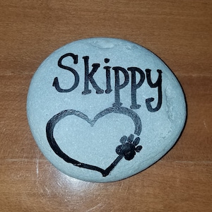 May include: A gray painted rock with the name "Skippy" written in black paint. A black heart with a paw print inside is painted on the rock.