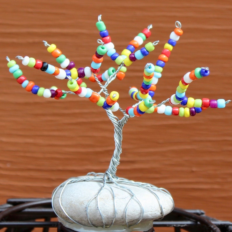 Pode incluir: Uma escultura de &aacute;rvore de arame com contas coloridas com uma base de pedra branca. A &aacute;rvore &eacute; feita de arame e contas de v&aacute;rias cores, incluindo vermelho, laranja, amarelo, verde, azul e roxo.