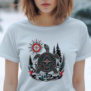 May include: A light gray t-shirt featuring a graphic print of a turtle with intricate geometric patterns in red, teal, and black. The design includes a stylized sun, evergreen trees, and rocky terrain elements surrounding the central turtle figure.