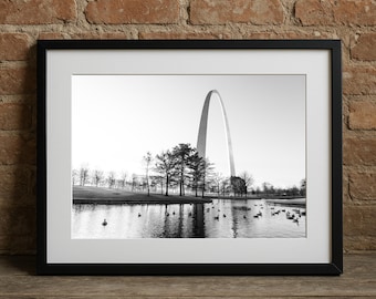 May include: Black and white framed photograph of the Gateway Arch in St. Louis, Missouri. The arch is reflected in the water of a pond, with trees and ducks in the foreground. The photograph is in a black frame.