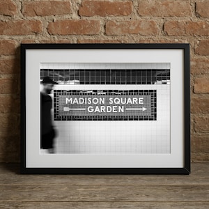 May include: Black and white framed print featuring a subway sign that reads "MADISON SQUARE GARDEN" with an arrow. The image is set against a backdrop of white and black tiles, with a blurred figure in the background.