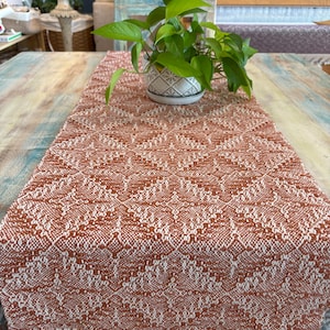 May include: A woven table runner in shades of rust and cream, featuring a geometric pattern and fringed edges. A potted plant with green leaves sits on the runner. The runner is displayed on a wooden table.